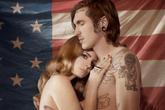 Topless Lana del Rey embraced by a tattooed man in front of the American flag.